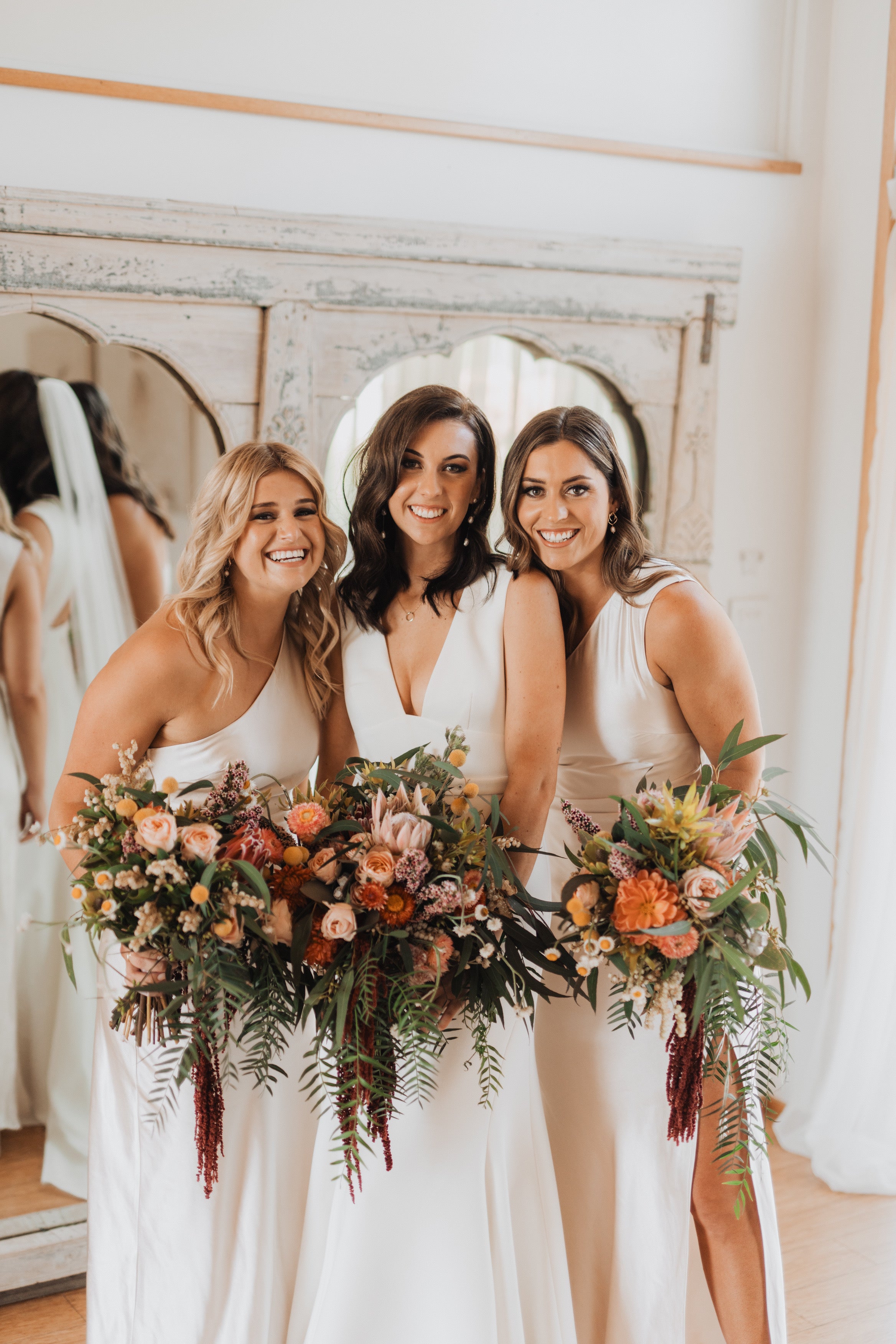 This vibrant image features a bride and her two bridesmaids, all dressed in elegant ivory gowns, posing together with radiant smiles. They each hold lush, colorful bouquets filled with a mix of fresh flowers and greenery, set against a beautifully rustic mirrored backdrop, creating a warm and joyful bridal moment.