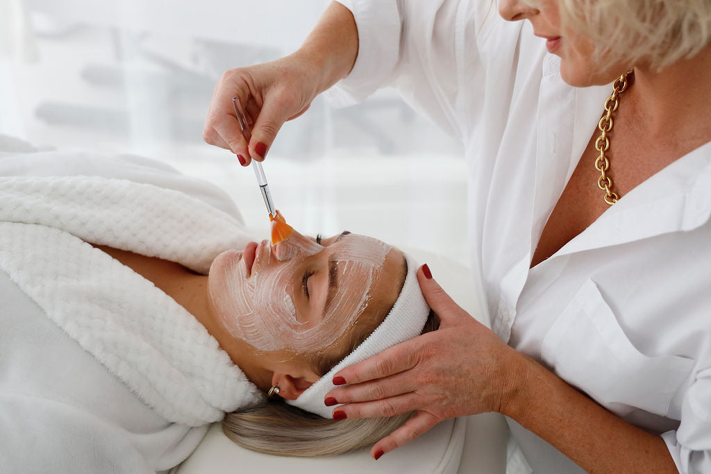 This image features a professional beautician applying a facial mask to a reclining client using a soft brush. The client is draped in a plush white robe and headband, while the bright, serene setting emphasizes relaxation and luxurious skincare treatment.