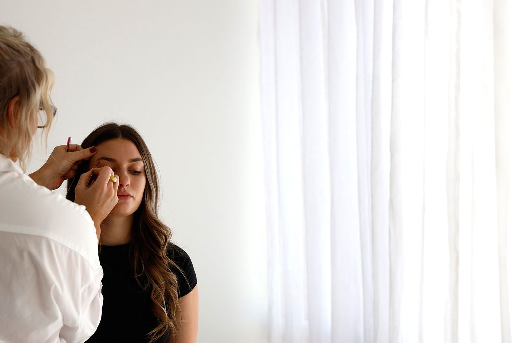 This image captures a beautician carefully applying makeup to a client in a serene, well-lit studio. The client sits calmly with her eyes closed as the professional works with precision, creating a peaceful atmosphere that emphasizes relaxation and expert service.