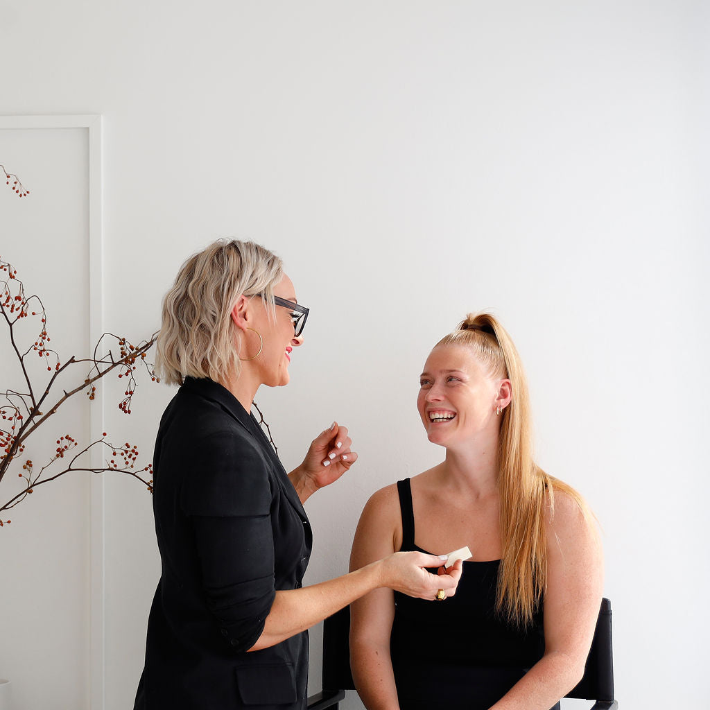 This image depicts a cheerful interaction between a professional makeup artist and her client during a beauty session. The client sits in a chair smiling warmly while the artist, dressed stylishly in black, prepares to apply a product in a bright, minimalist studio accented with delicate branches.