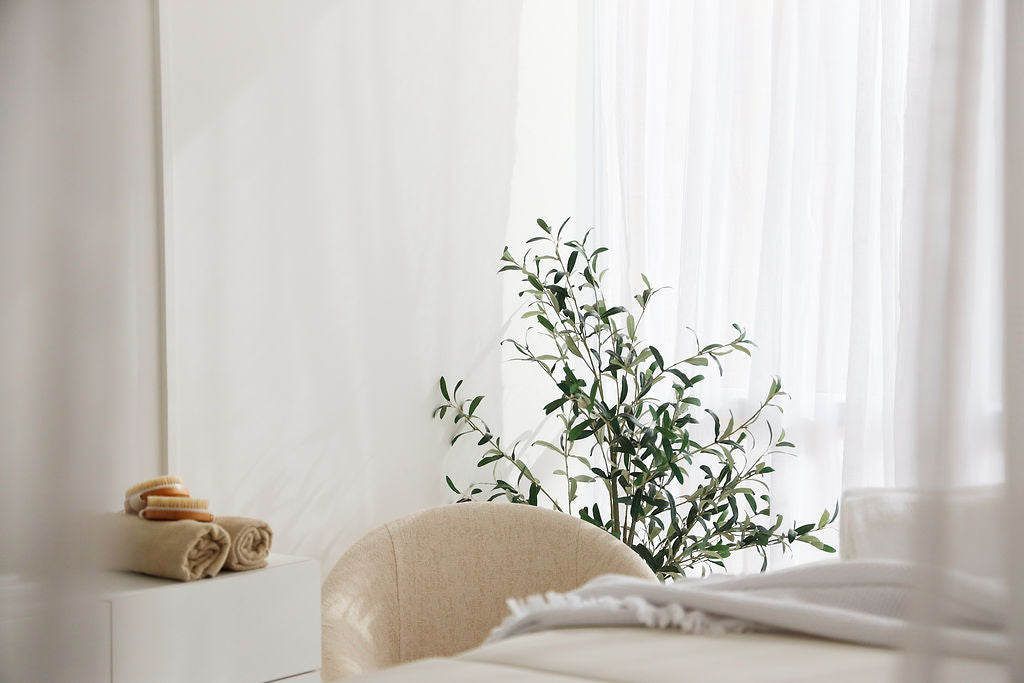 This image shows a serene, minimalist interior featuring a beige chair, neatly folded towels, and a small stack of soaps on a table. A leafy green plant adds a natural touch against a backdrop of soft, white curtains that allow diffused natural light to fill the room. Perfect for evoking a calm, spa-like ambiance.
