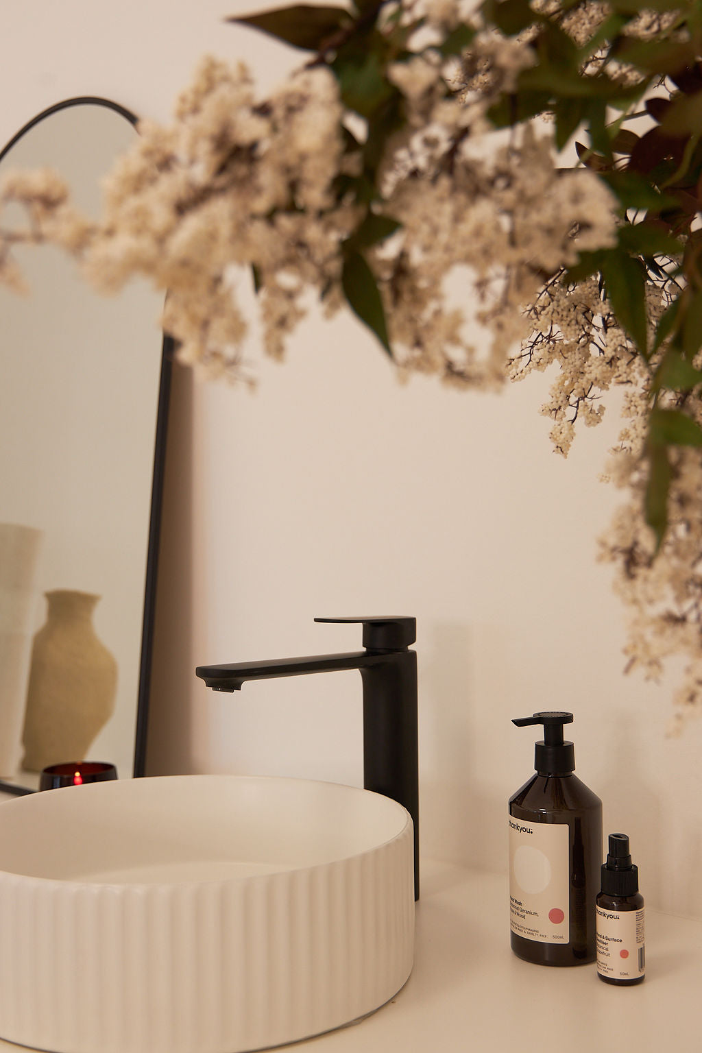 This image showcases a stylish spa or skincare studio setup, featuring a modern ribbed white ceramic sink with a sleek black faucet. Nearby, skincare products in minimalist packaging are neatly arranged, complemented by soft floral accents and a reflective mirror, creating a serene and inviting ambiance.