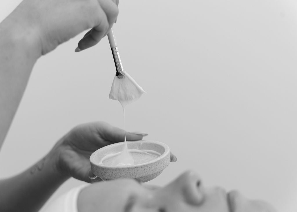 This black-and-white close-up image showcases a beautician preparing to apply a creamy facial mask using a soft fan brush. The product drips delicately from the brush into a small ceramic bowl, capturing the essence of a luxurious hydrating facial treatment.