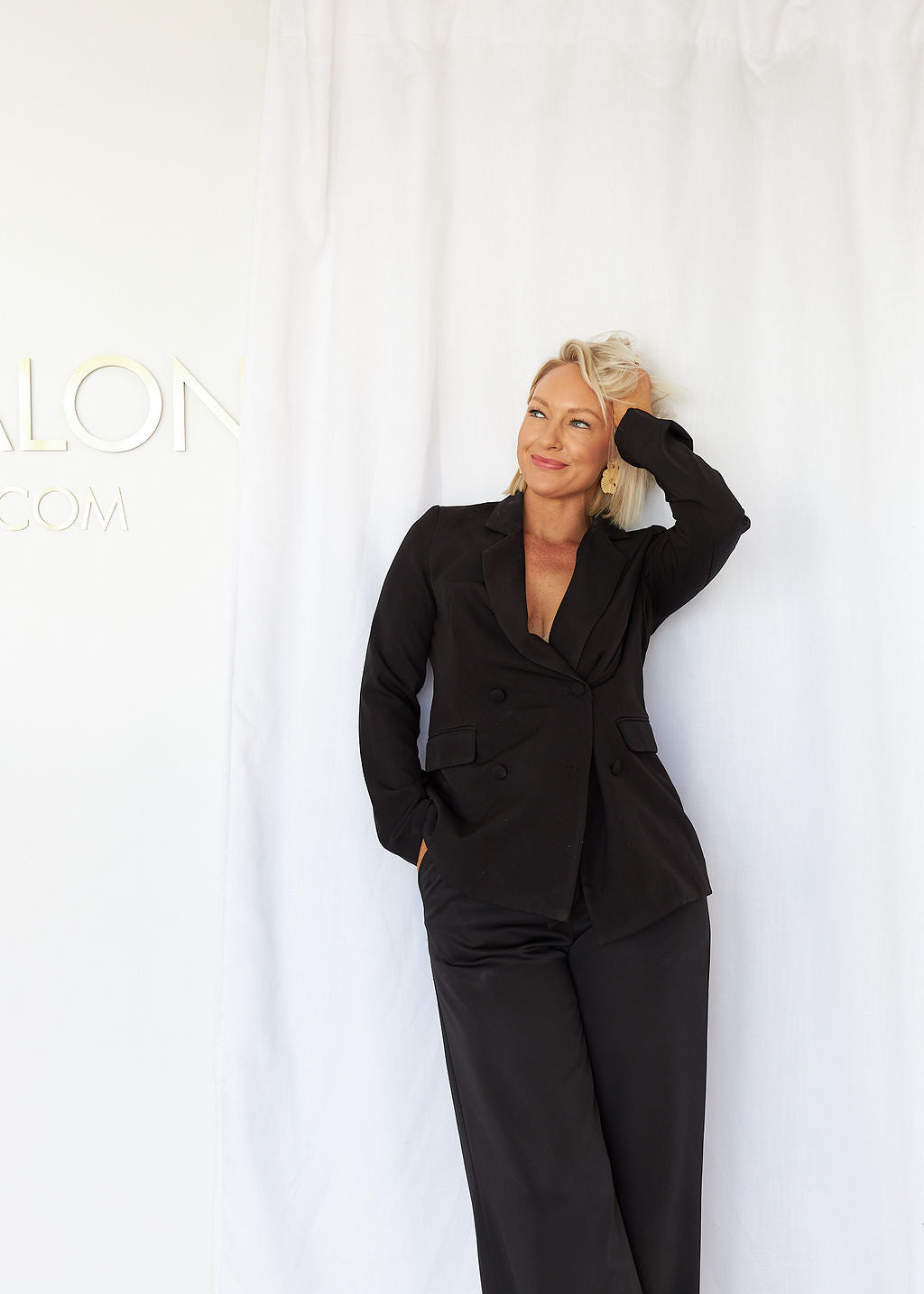 This image features a confident woman posing in a tailored blazer, leaning casually against a white backdrop. Her hand rests playfully in her hair, and subtle text partially visible in the background suggests a professional setting or brand representation.