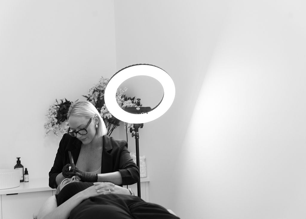This black-and-white image features a beautician focused on performing a precision facial treatment, illuminated by a professional ring light. The calm, minimalist studio includes floral arrangements and skincare products in the background, emphasizing a high-end and luxurious self-care experience.
