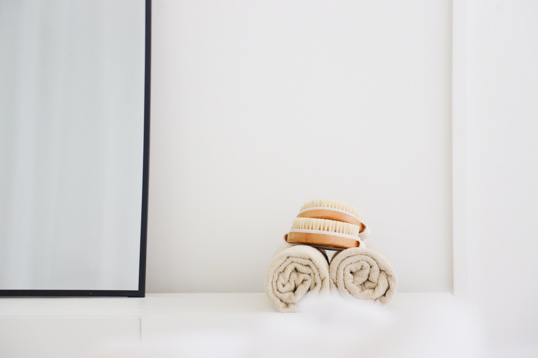 This minimalist image showcases a serene spa setup with three neatly rolled beige towels topped by two natural-bristle dry brushes with wooden handles. The composition conveys simplicity, cleanliness, and relaxation, emphasizing a wellness or self-care theme.
