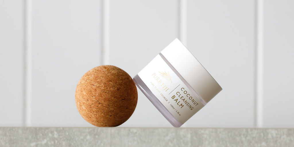 This minimalist image highlights a jar of "Pure Fiji Coconut Cleansing Balm" elegantly balanced on a natural cork sphere. The clean and modern backdrop emphasizes the product's simplicity and natural essence, reflecting its premium and organic skincare focus.