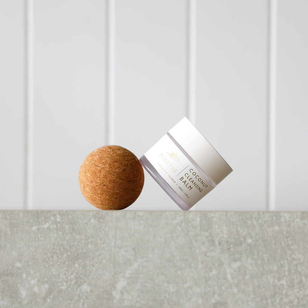 This minimalist image highlights a jar of "Pure Fiji Coconut Cleansing Balm" elegantly balanced on a natural cork sphere. The clean and modern backdrop emphasizes the product's simplicity and natural essence, reflecting its premium and organic skincare focus.