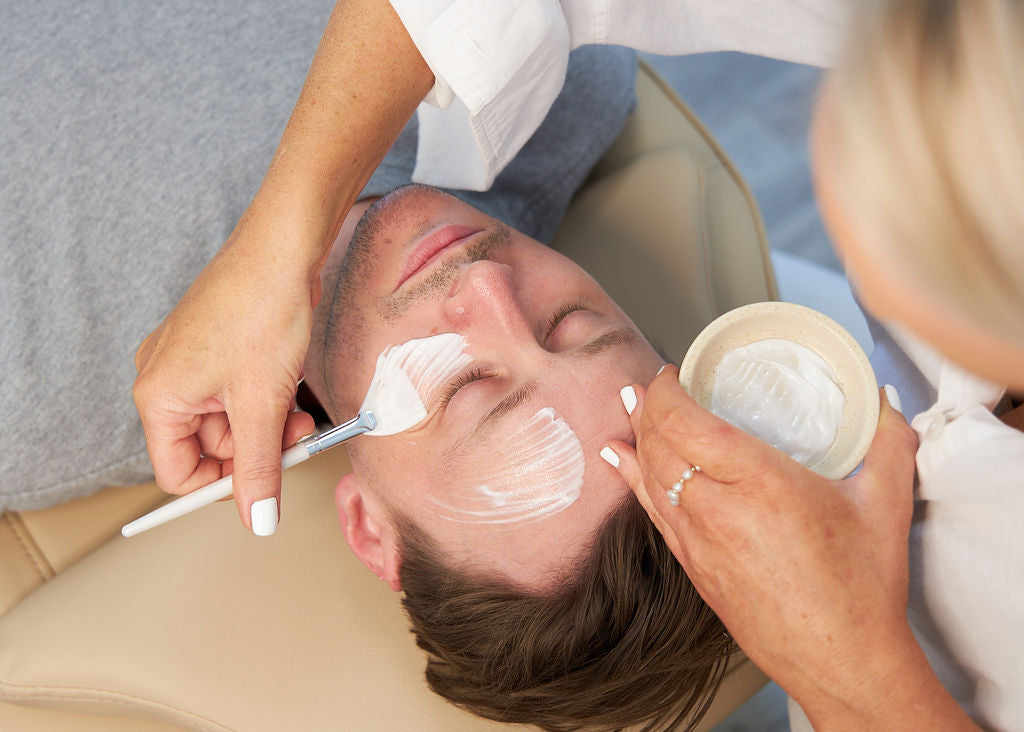 This image shows a relaxing facial treatment being performed on a male client. A beautician carefully applies a creamy mask with a soft brush while the client rests peacefully on a cushioned surface, highlighting the soothing and inclusive nature of the spa experience.